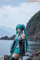 A woman with long blue hair standing on a rocky beach.