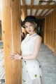 A woman in a white dress and black hat leaning against a wooden pole.